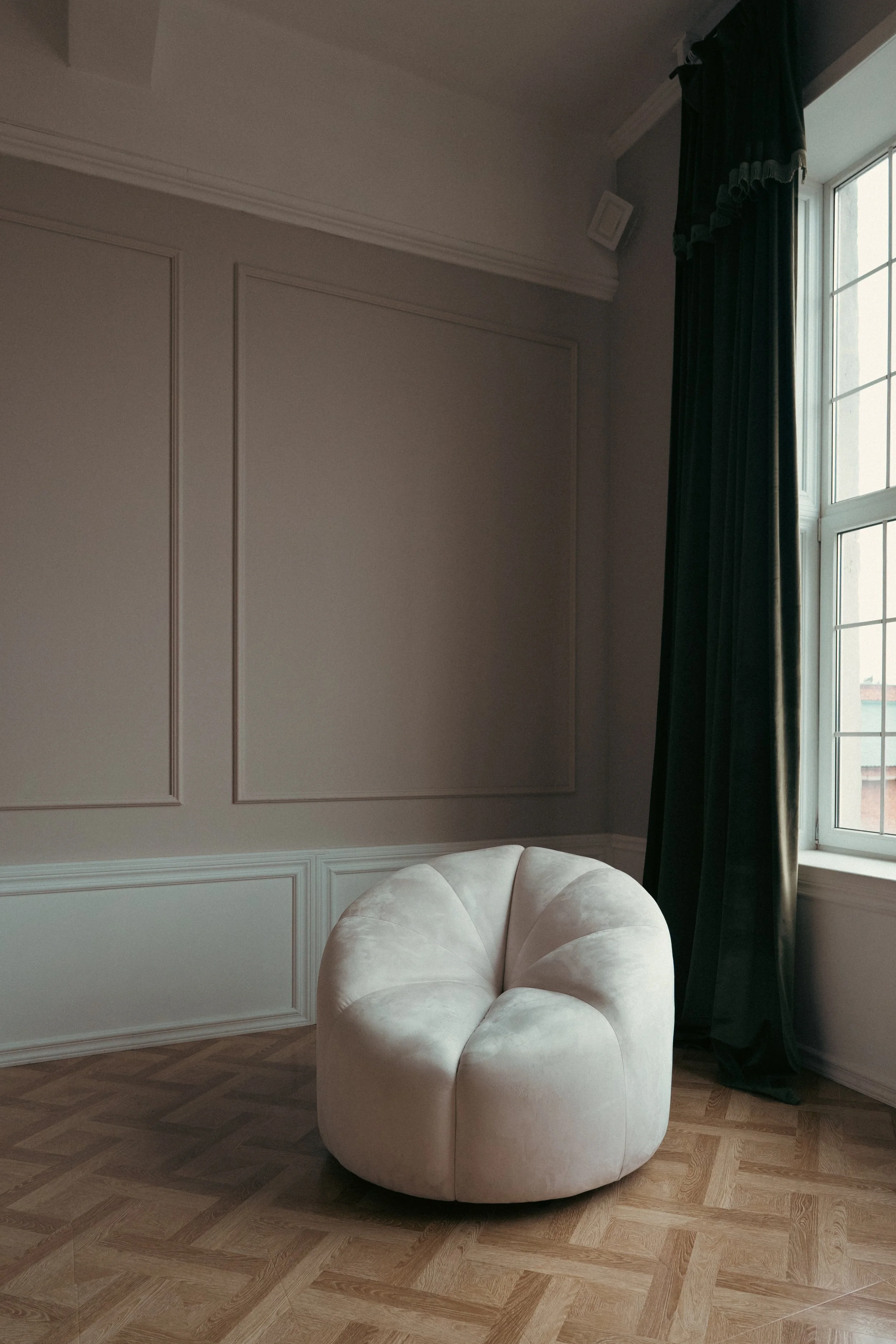 A cream-colored, upholstered, velvet armchair with rounded edges and button-tufted design in a room with wooden parquet flooring, a white wall with decorative molding, and a window with dark curtains.