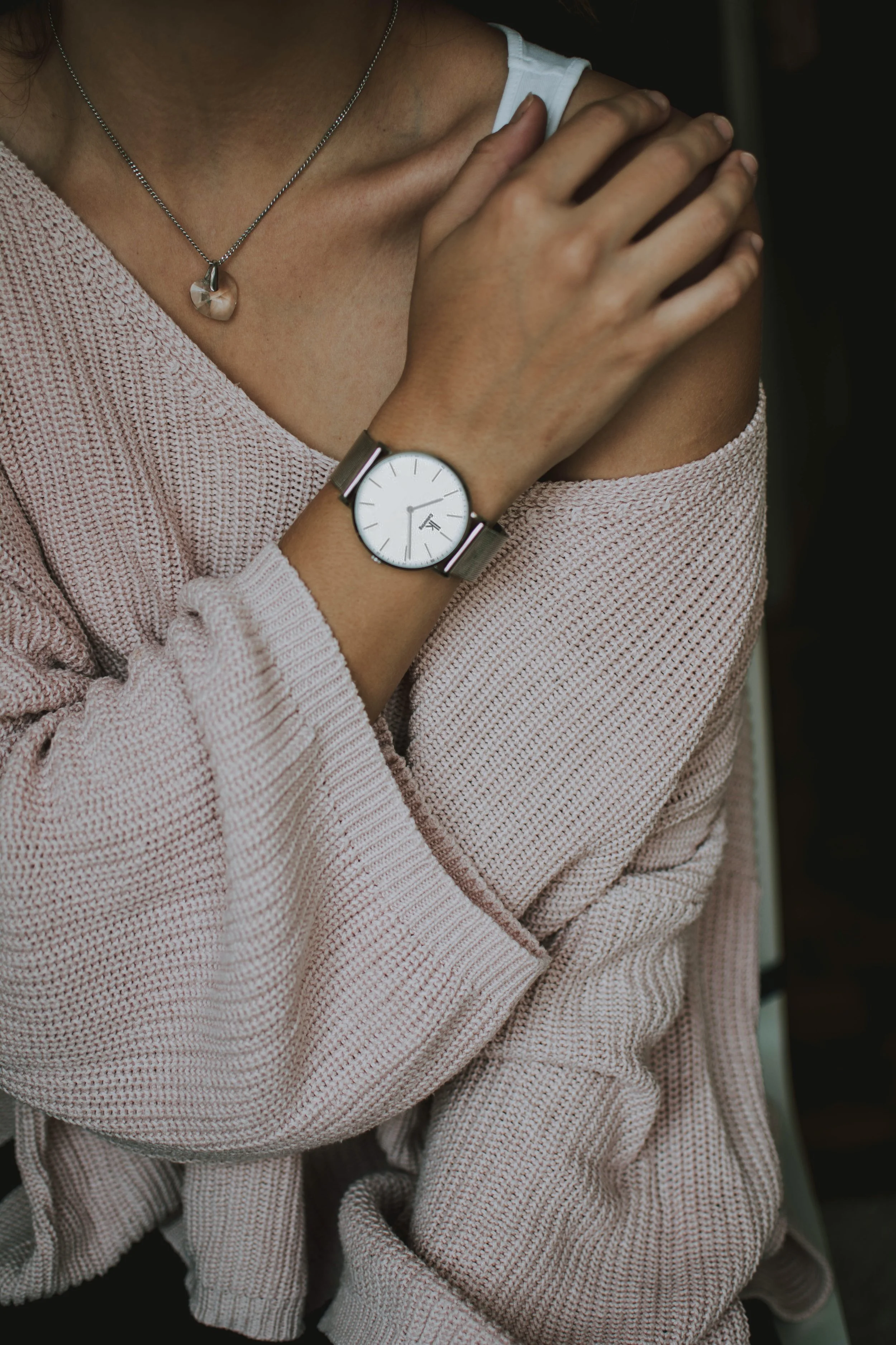 Close-up of a woman's cross-armed pose showing a silver wristwatch and a delicate necklace with a heart-shaped pendant, wearing a light pink Knit sweater and a white tank top underneath.