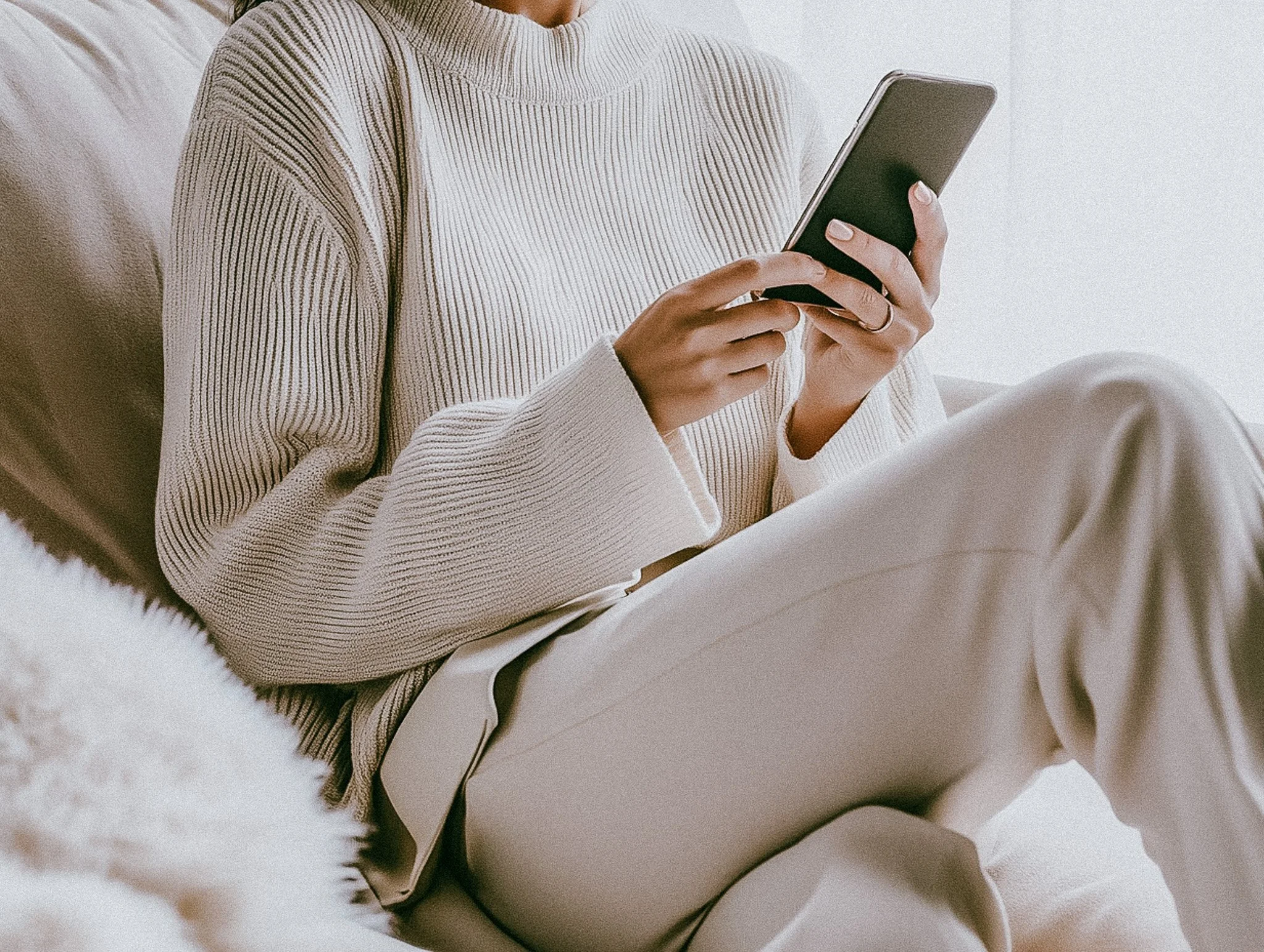 A person wearing a beige ribbed sweater and light-colored pants, sitting on a white surface and holding a smartphone.