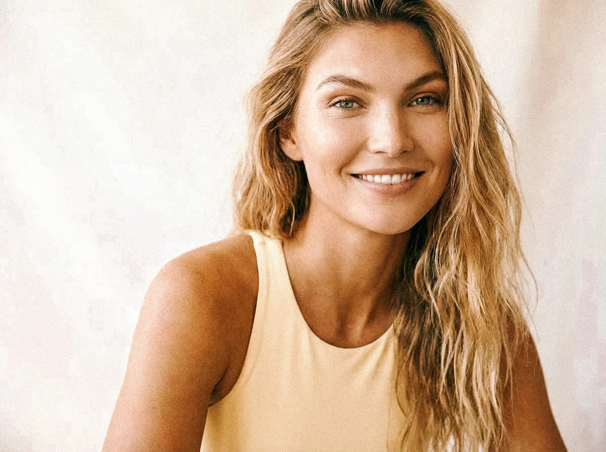 A woman with long, wavy blonde hair smiling, wearing a light-colored sleeveless top, against a neutral background.