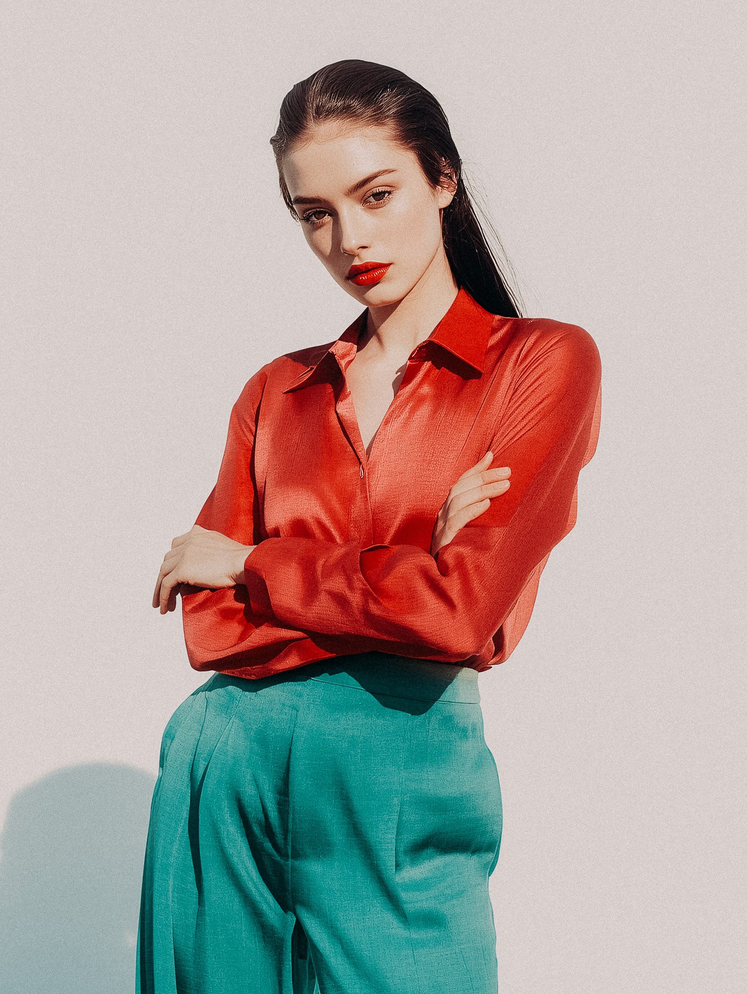 A woman with wet black hair, red lipstick, wearing a red blouse and teal pants, standing with arms crossed on a plain light background.
