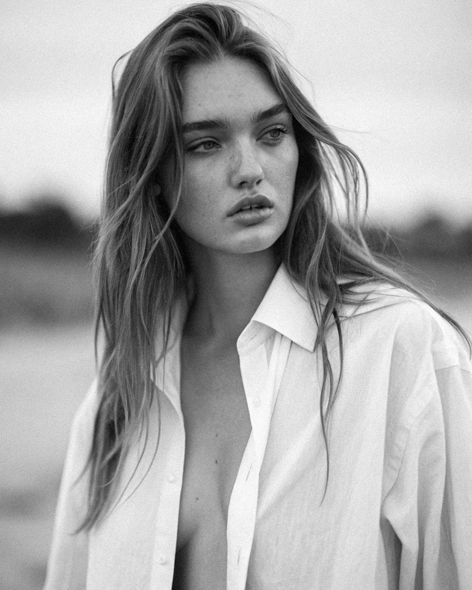 A black and white photo of a woman with long hair, wearing a white shirt, looking thoughtfully into the distance.