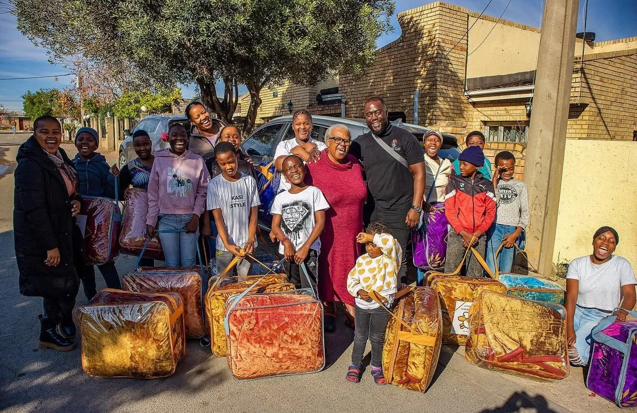 A group of smiling children and adults standing on a street, holding colorful suitcases and bags, beside a parked car and a tree in a neighborhood with brick buildings.