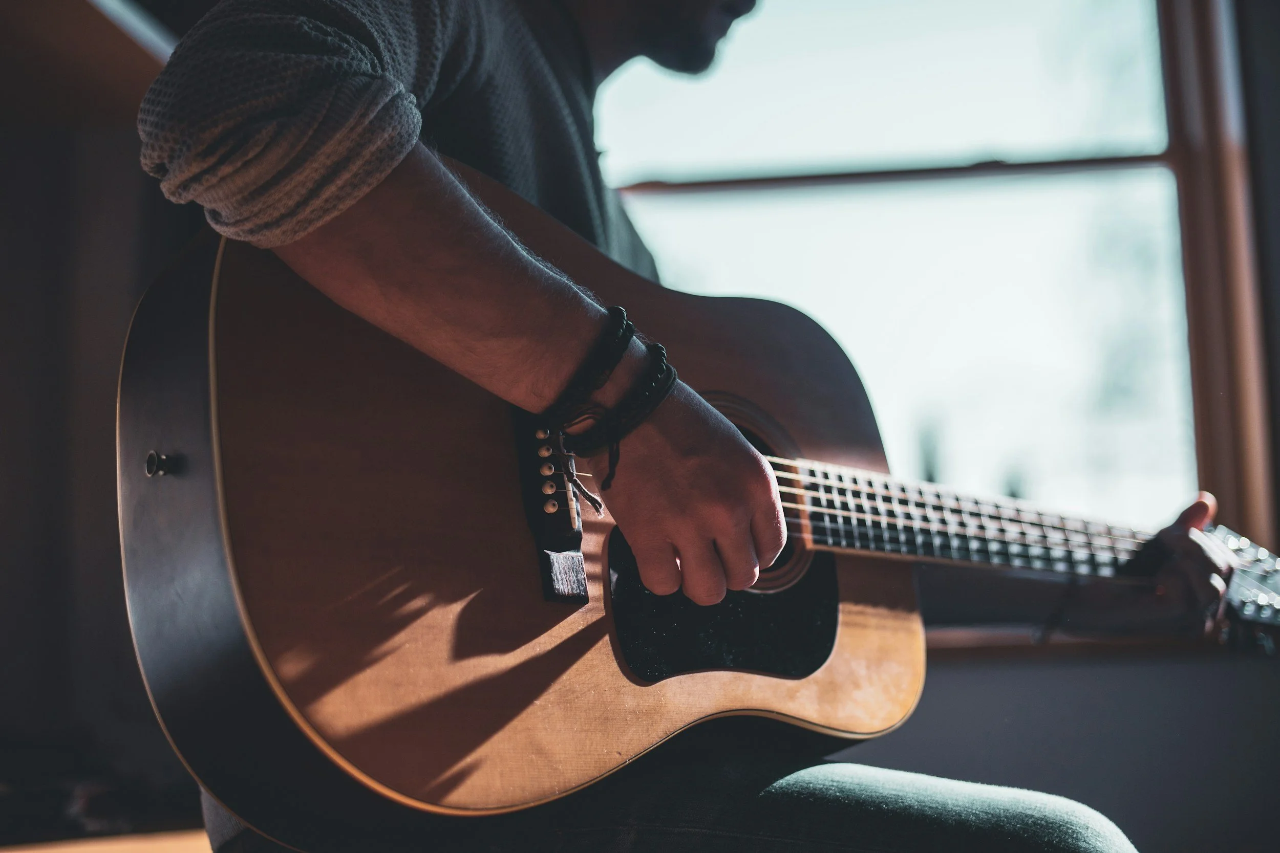 A person playing an acoustic guitar with their right arm and hand strumming near the strings, wearing a textured gray sleeve and black bracelets, indoors near a window with light coming through.