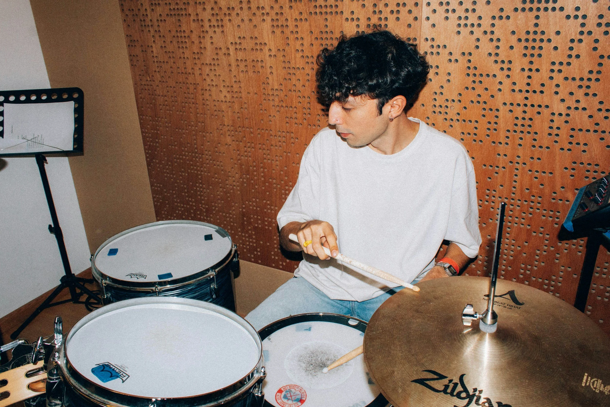 A young man with dark curly hair and earrings playing drums in a music studio with wooden soundproofing walls, surrounded by drums and cymbals.