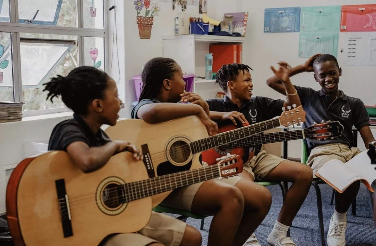 Four school children sitting in a classroom, two holding guitars, smiling and sharing a moment of laughter.