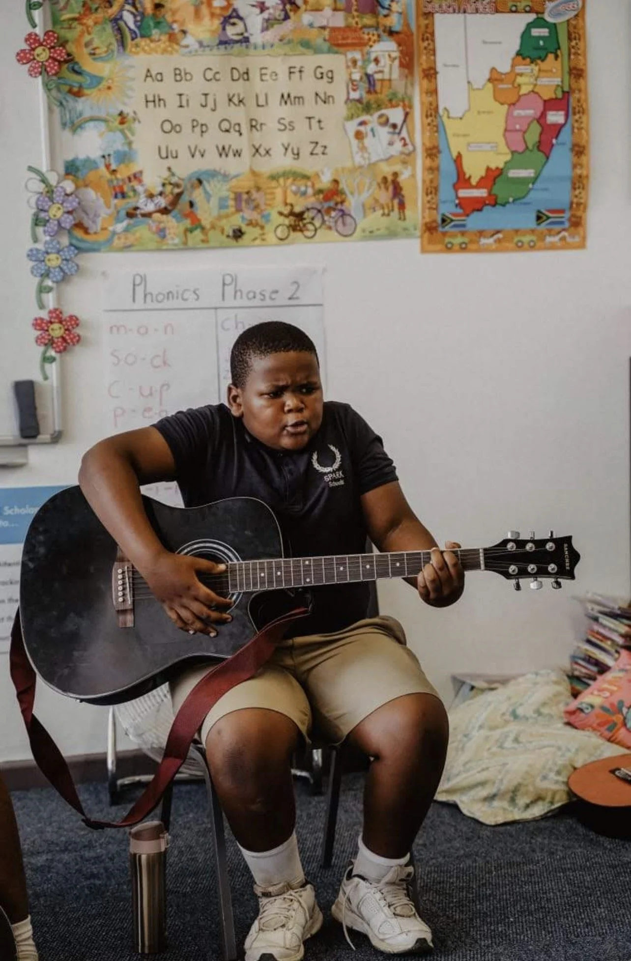 A young boy sitting on a chair playing an acoustic guitar in a classroom. The boy is wearing a black shirt, khaki shorts, white socks, and white sneakers. The classroom has educational posters, a map of South Africa, and colorful decorations on the whiteboard and wall.