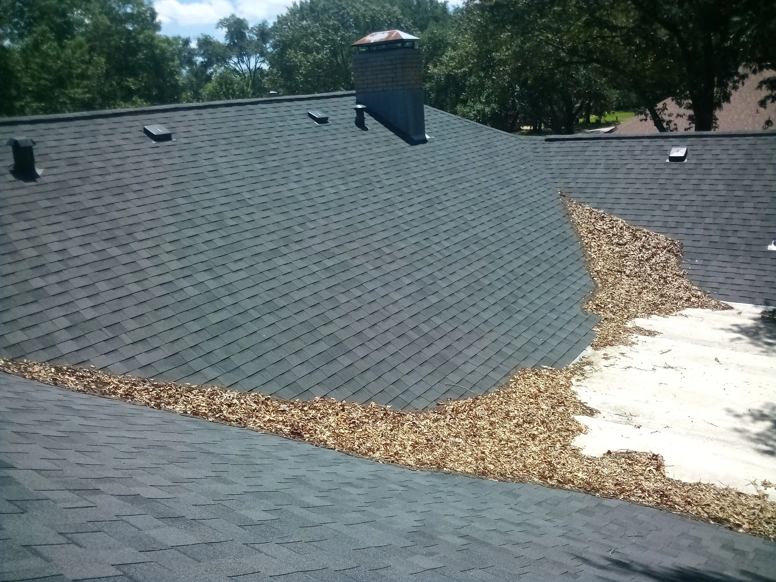 A rooftop with gray shingles, four vents, a brick chimney, and a pile of brown mulch on the side.