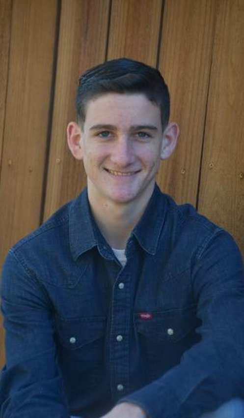 Young man with short dark hair and blue eyes smiling, wearing a dark denim button-up shirt, sitting in front of a wooden fence.