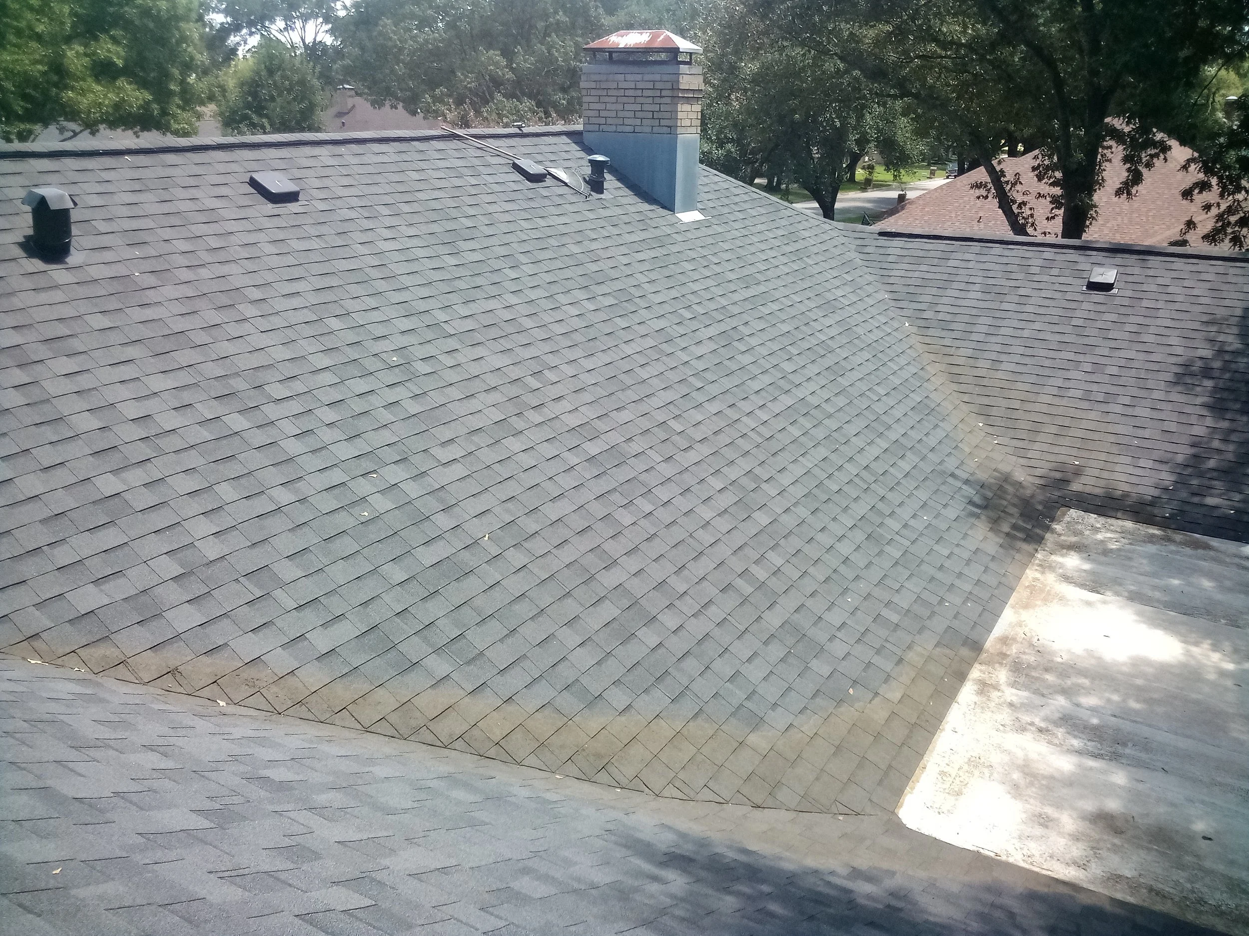 A residential house roof covered in grey asphalt shingles, with a chimney, ventilation pipes, and a tree with green leaves in the background.