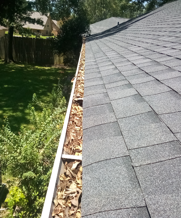 View of a roof with new gray shingles and an existing gutter with leaves, next to a yard with green grass, bushes, and trees.