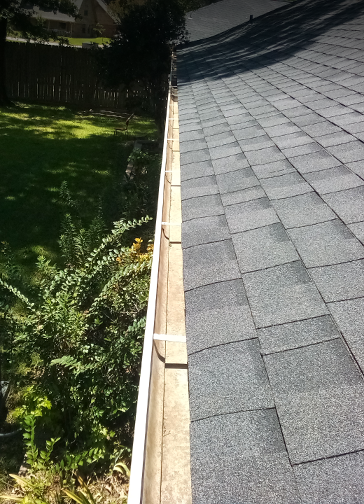 View of a house roof with gray shingles and a gutter, with a backyard on the left side containing green grass, bushes, and a wooden fence.