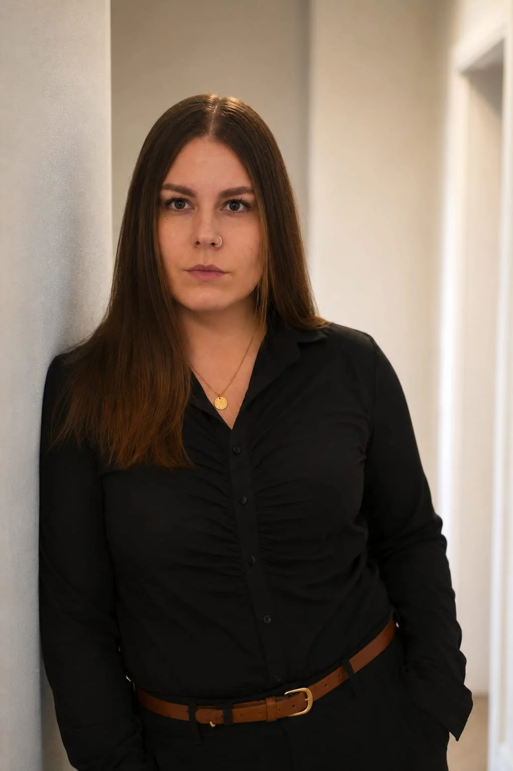 A young woman with long brown hair wearing a black blouse, standing against a light-colored wall in a hallway, looking directly at the camera.