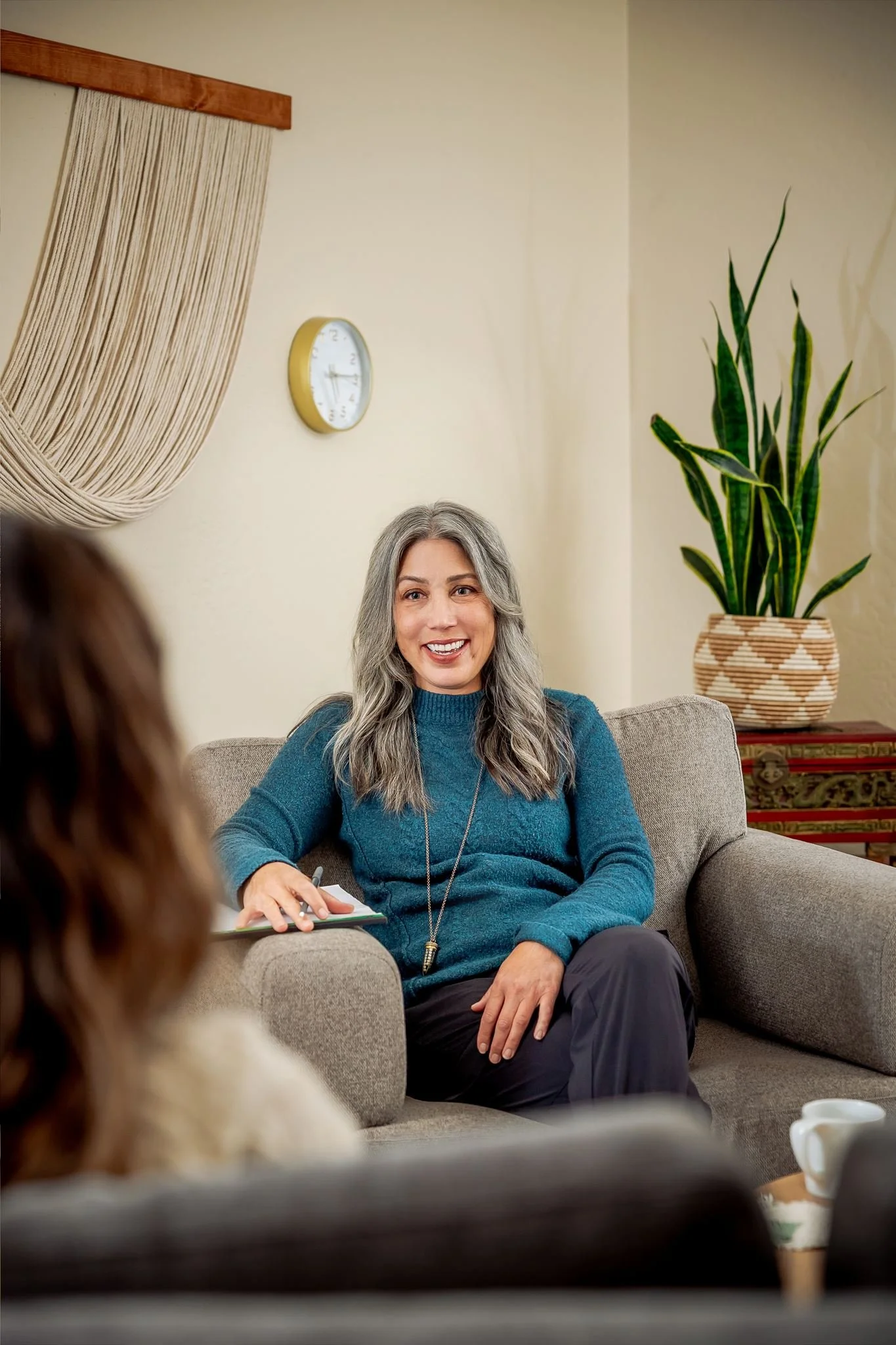 A woman with long gray hair, wearing a teal sweater, sitting on a beige couch, smiling, holding a notepad with a pen, engaged in a conversation with another person in a cozy room with a large green plant in a decorative basket, a clock on the wall, a curtain, and a table with a coffee mug.