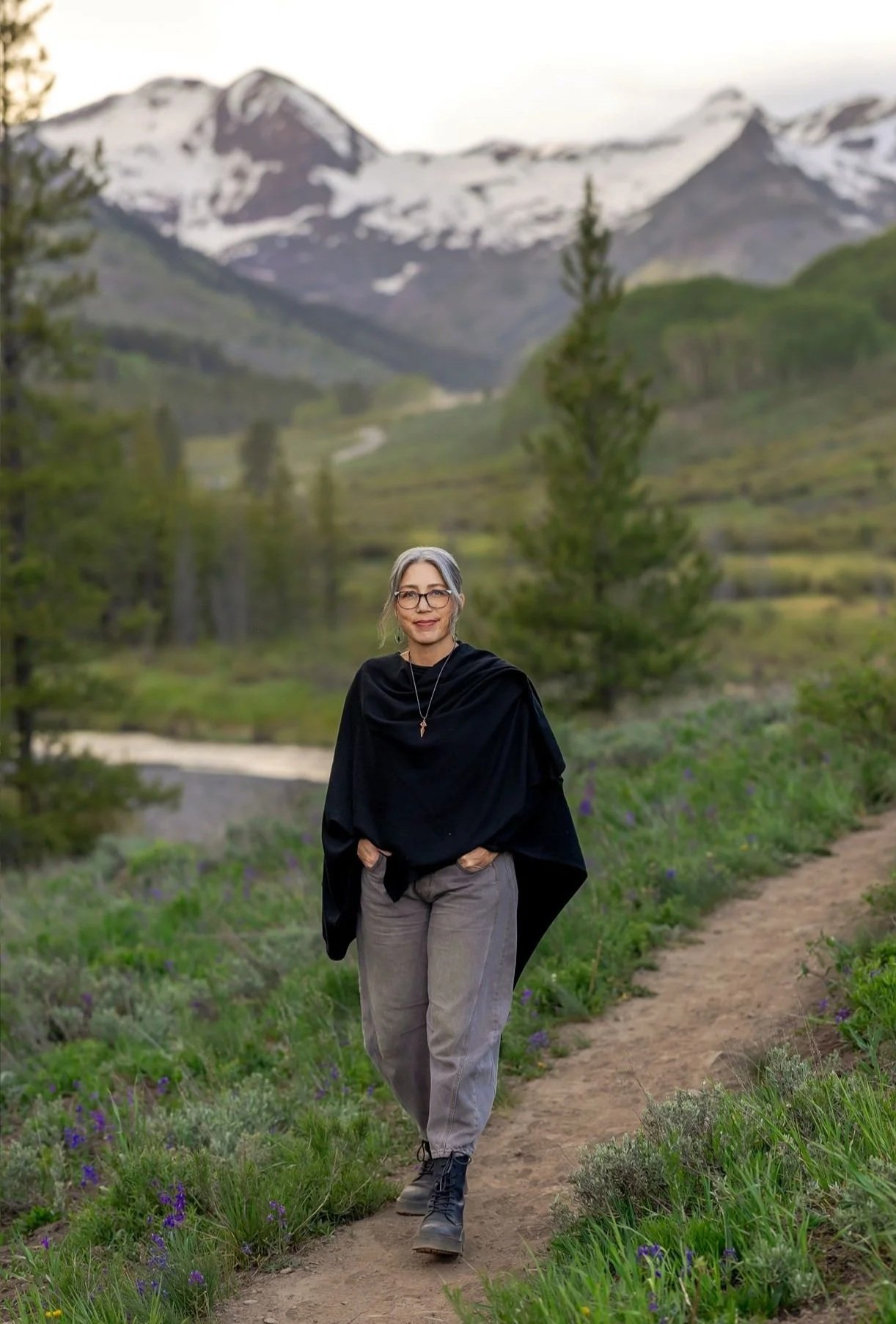 A woman with gray hair, glasses, and a black shawl walking on a dirt path through green grass and purple wildflowers with mountains and snow in the background.