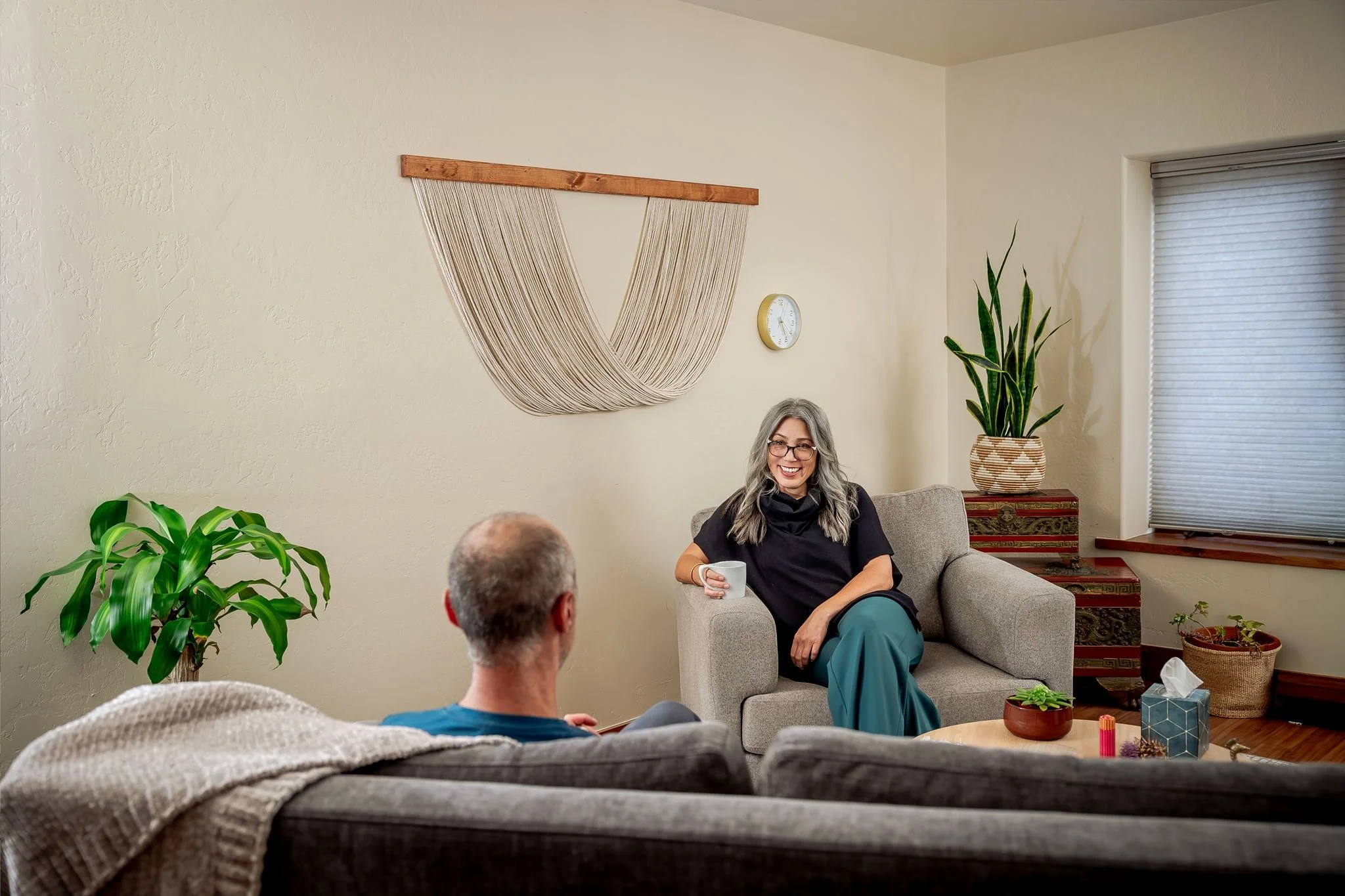A smiling woman with gray hair and glasses sitting on a beige sofa, holding a white mug, engaged in conversation with two people in a cozy living room decorated with plants, a wall clock, and woven wall hanging.