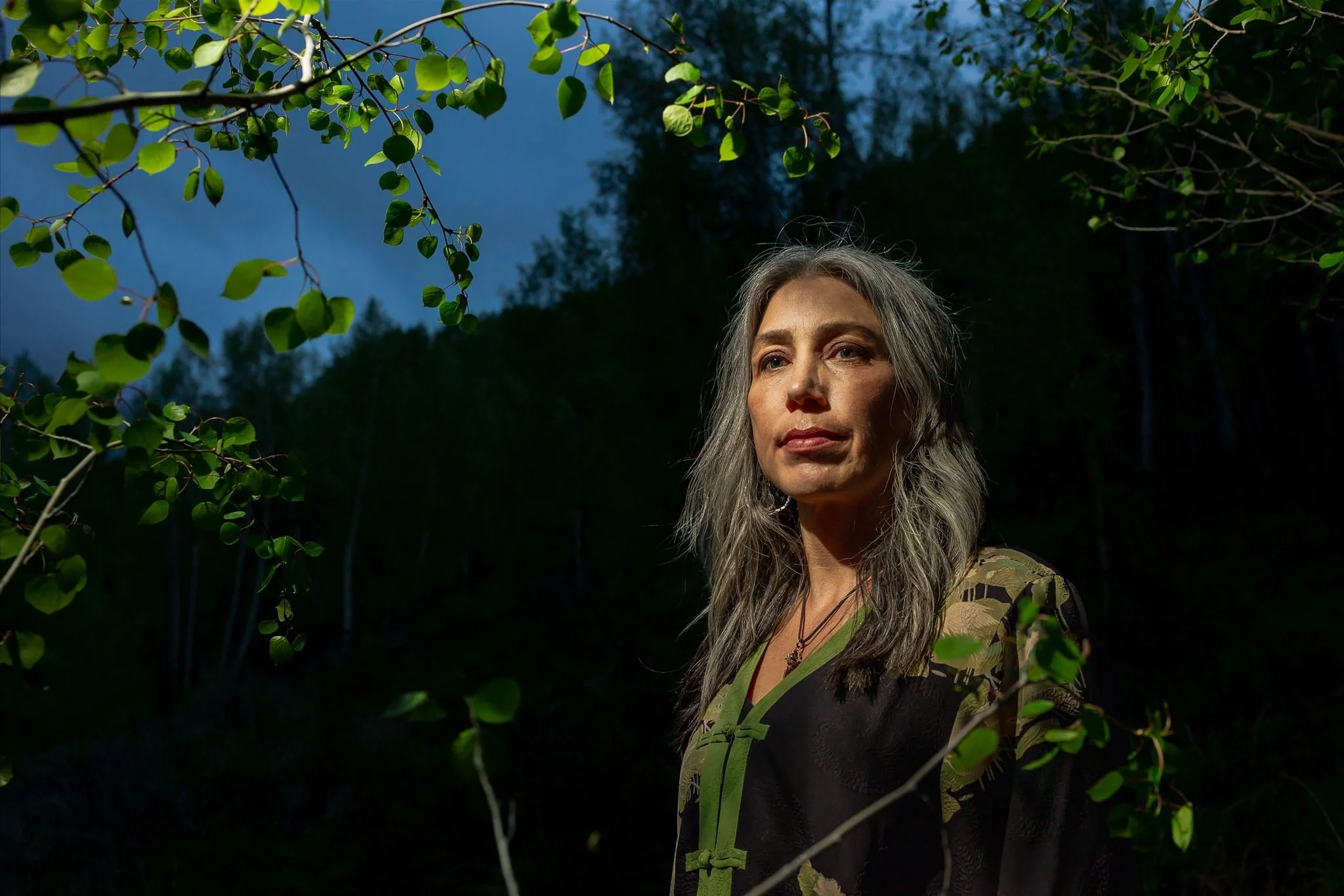 A woman with long gray hair standing outdoors among green leaves and trees during dusk or night, with a dark sky in the background.