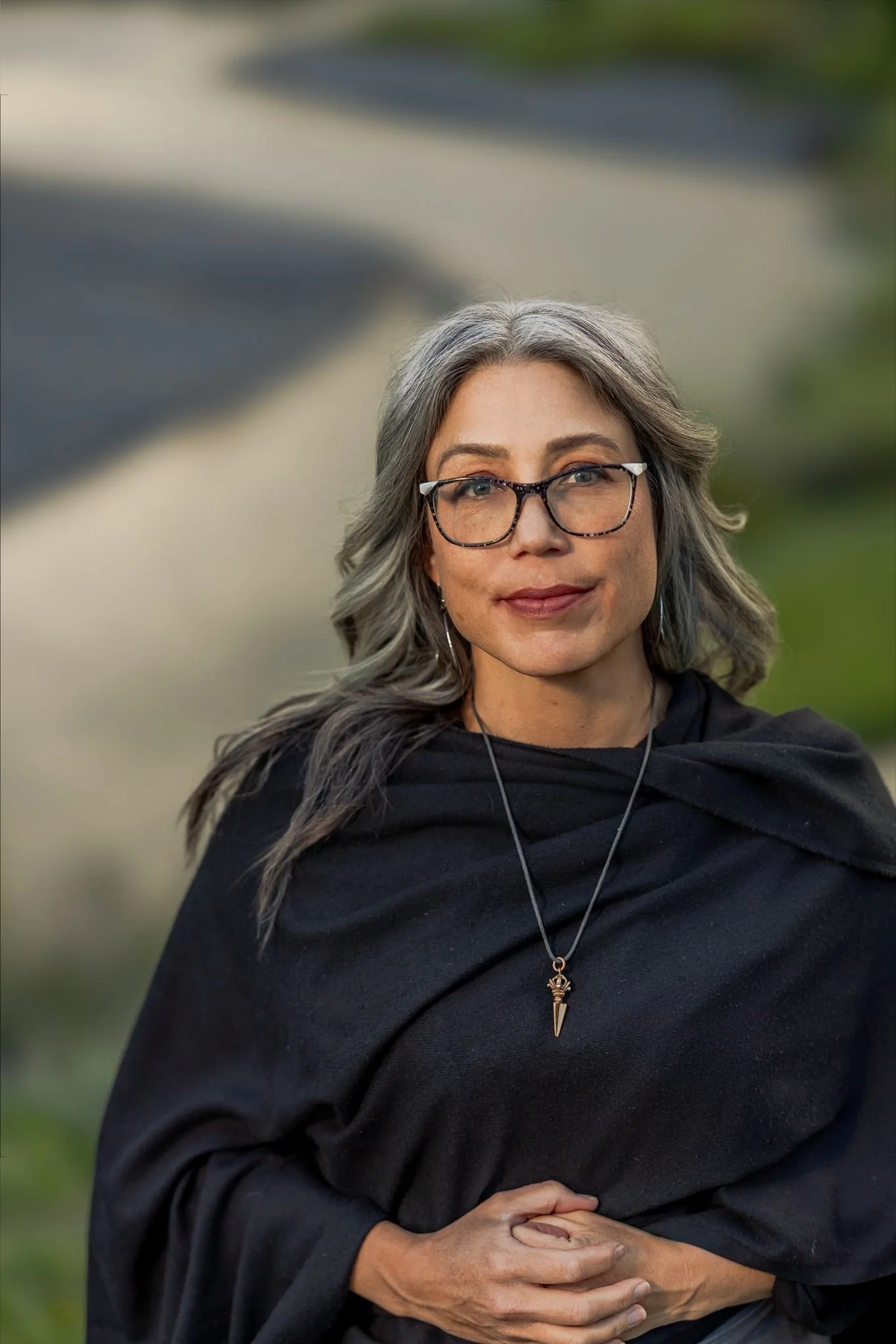 A woman with gray and black hair, wearing glasses, a black top, and a necklace with a pendant, stands outdoors with blurred greenery in the background. She has a calm expression and her hands are crossed and resting on her stomach.