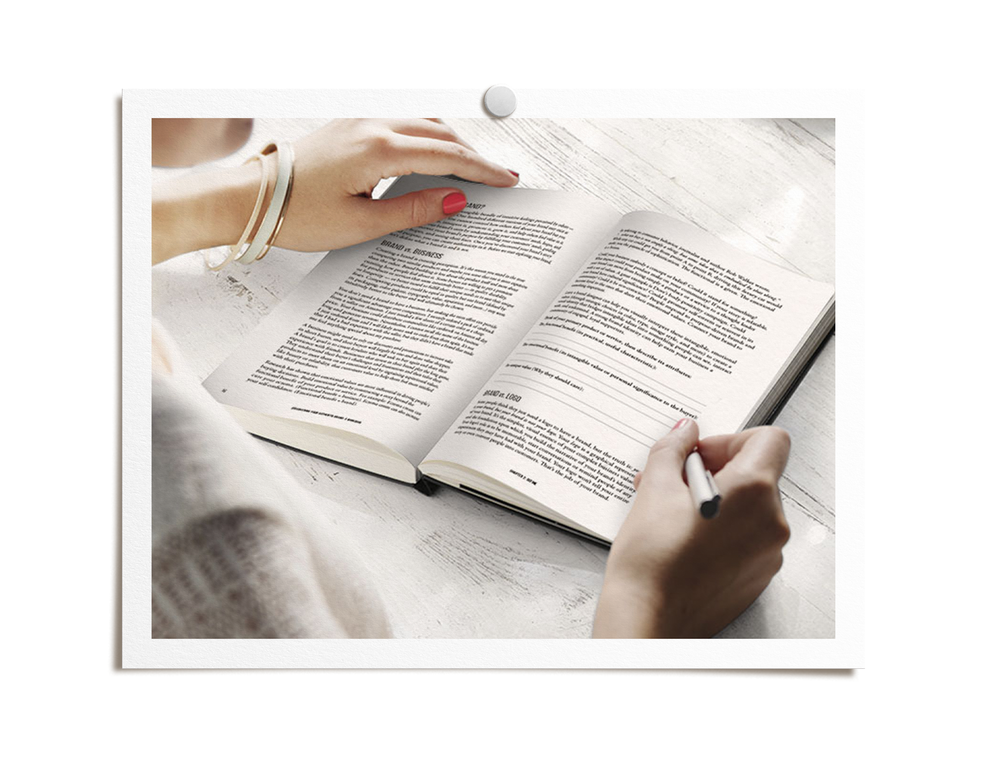 A person's hand with pink nail polish and light-colored bracelets is holding a page of a book. Another hand with a black pen is writing in the book. The book is open on a light-colored wooden surface.