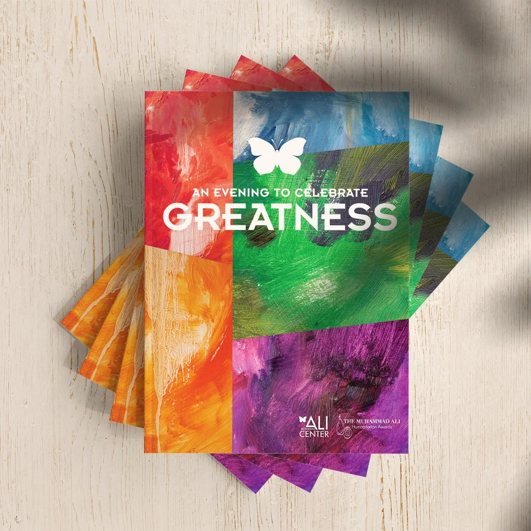 Colorful flyers for an event titled "An Evening to Celebrate Greatness" with a white butterfly logo, organized by the Ali Center and Muhammad Ali Humanitarian Awards, displayed on a wooden surface.