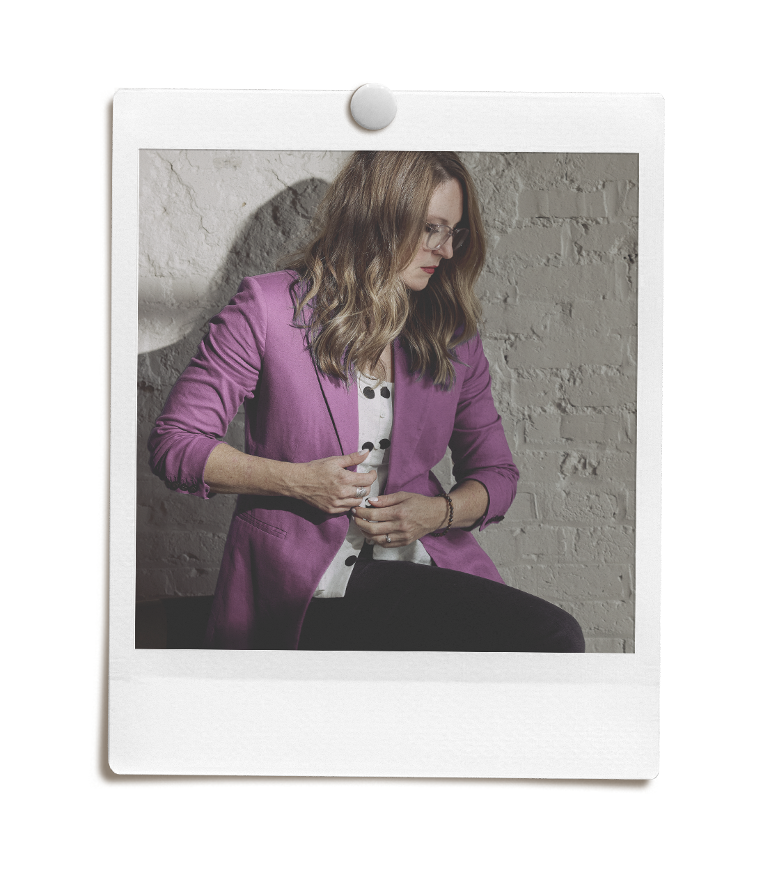 A woman with wavy blonde hair and glasses sitting against a brick wall, wearing a purple blazer and a polka dot shirt.