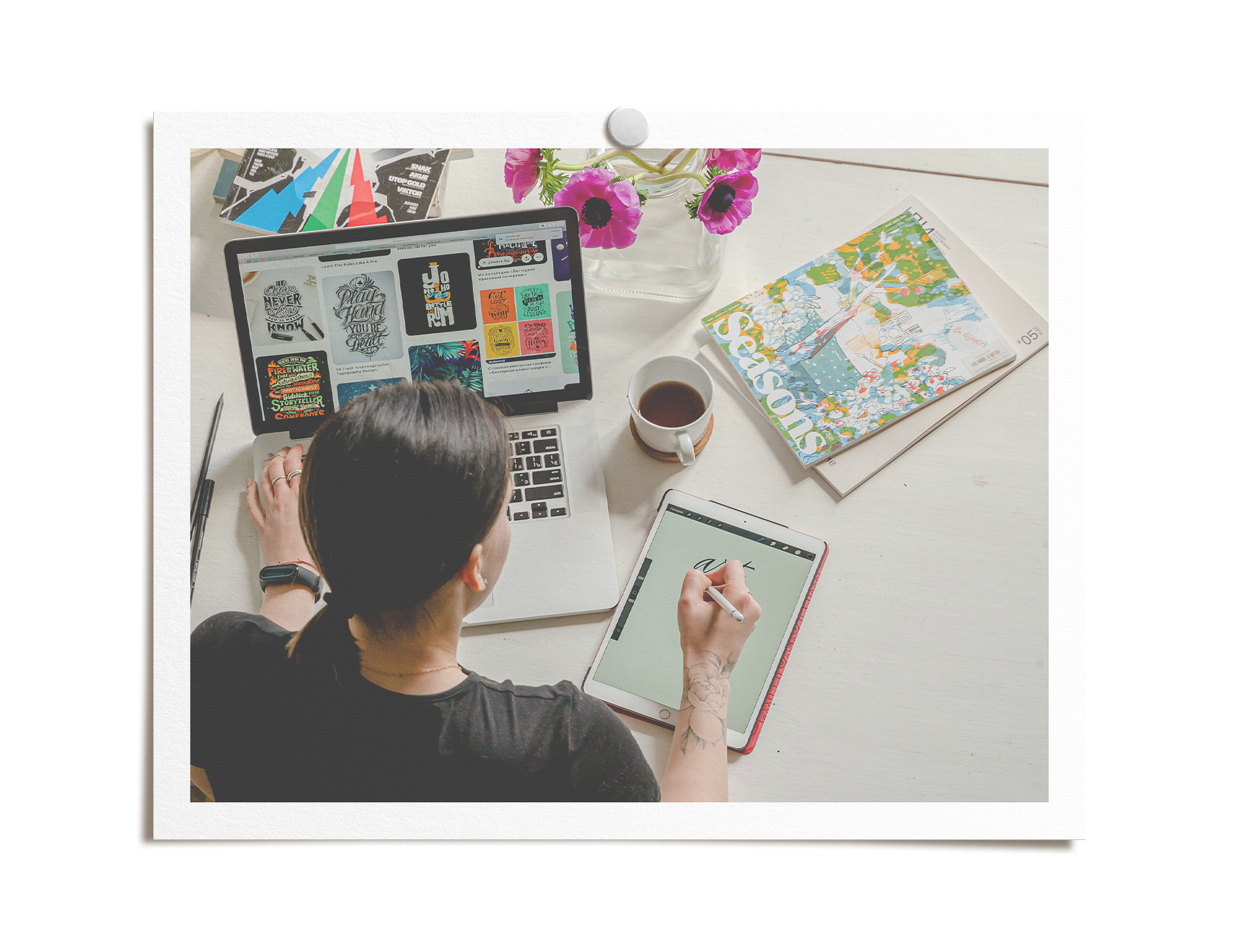 A graphic designer preparing for a new project. Various items are on the desk including a laptop displaying colorful designs, a tablet with a green screen, a cup of coffee, a vase with pink flowers, and colorful papers.