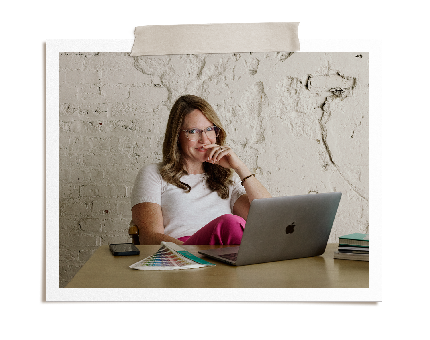 A woman with glasses sitting at a desk with a laptop, color swatches, and notebooks, smiling and looking at the camera, against a white brick wall background.