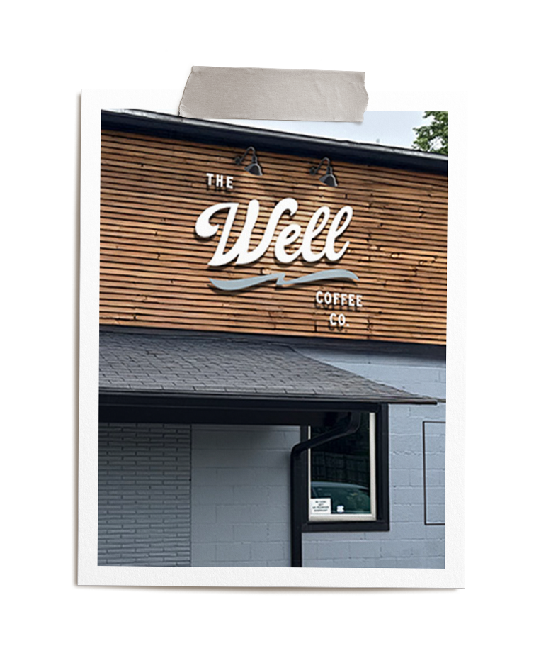 Sign for The Well Coffee Co. on a building with wooden and brick exterior, under a black roof with two spotlights.