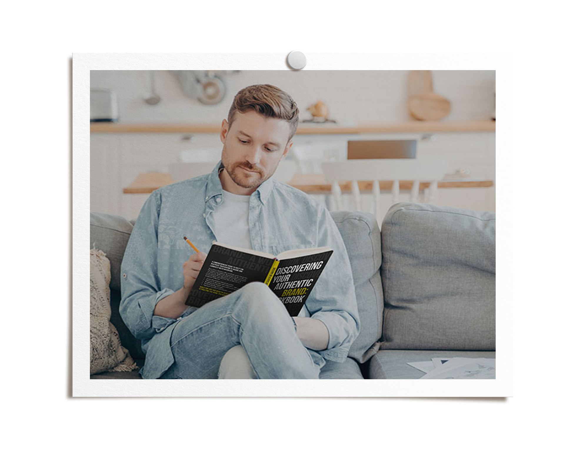 A young man in a light blue denim shirt and white t-shirt sitting on a beige sofa, reading a book titled 'Discovering Your Authentic Brand' in a bright, cozy living space.
