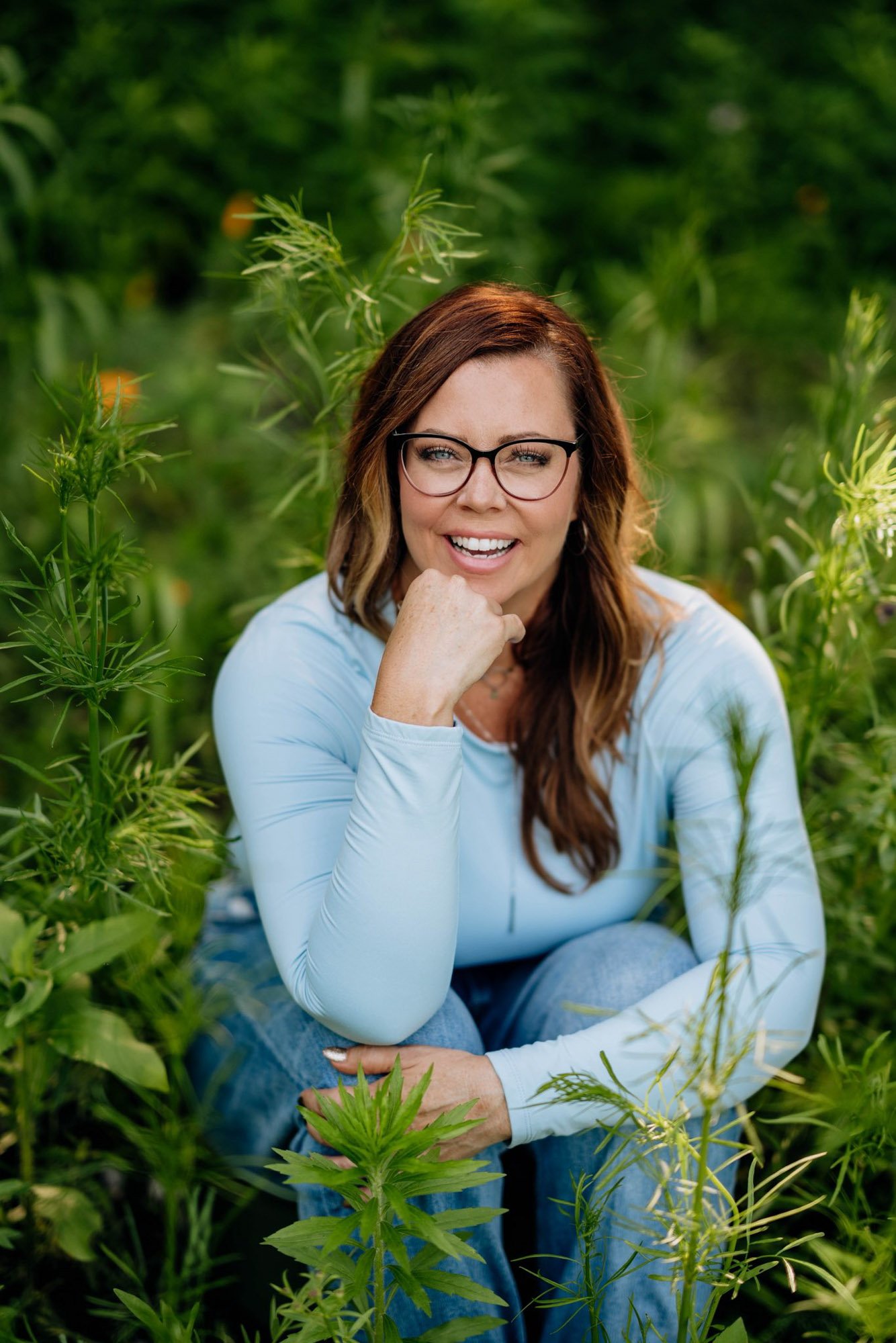 A woman with red hair, glasses, and a light blue long sleeve shirt sitting outdoors among green plants, smiling and resting her chin on her hand.