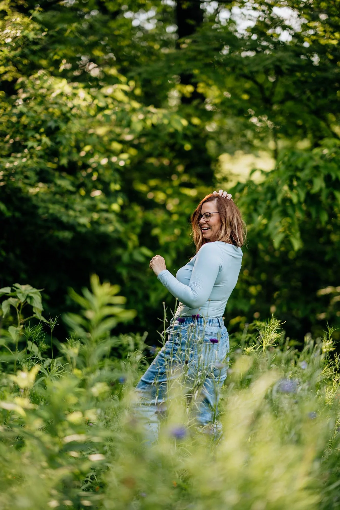 A woman with glasses, wearing a white long-sleeve shirt and denim skirt, smiling and standing in a lush green outdoor setting with dense foliage and tall plants.