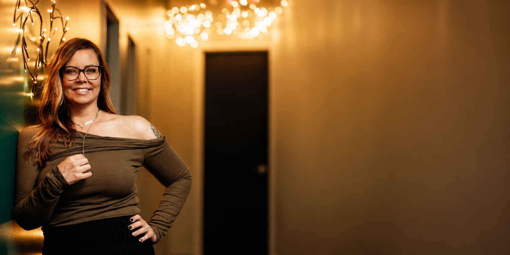 A woman with glasses and red hair, smiling, standing in a hallway decorated with string lights.