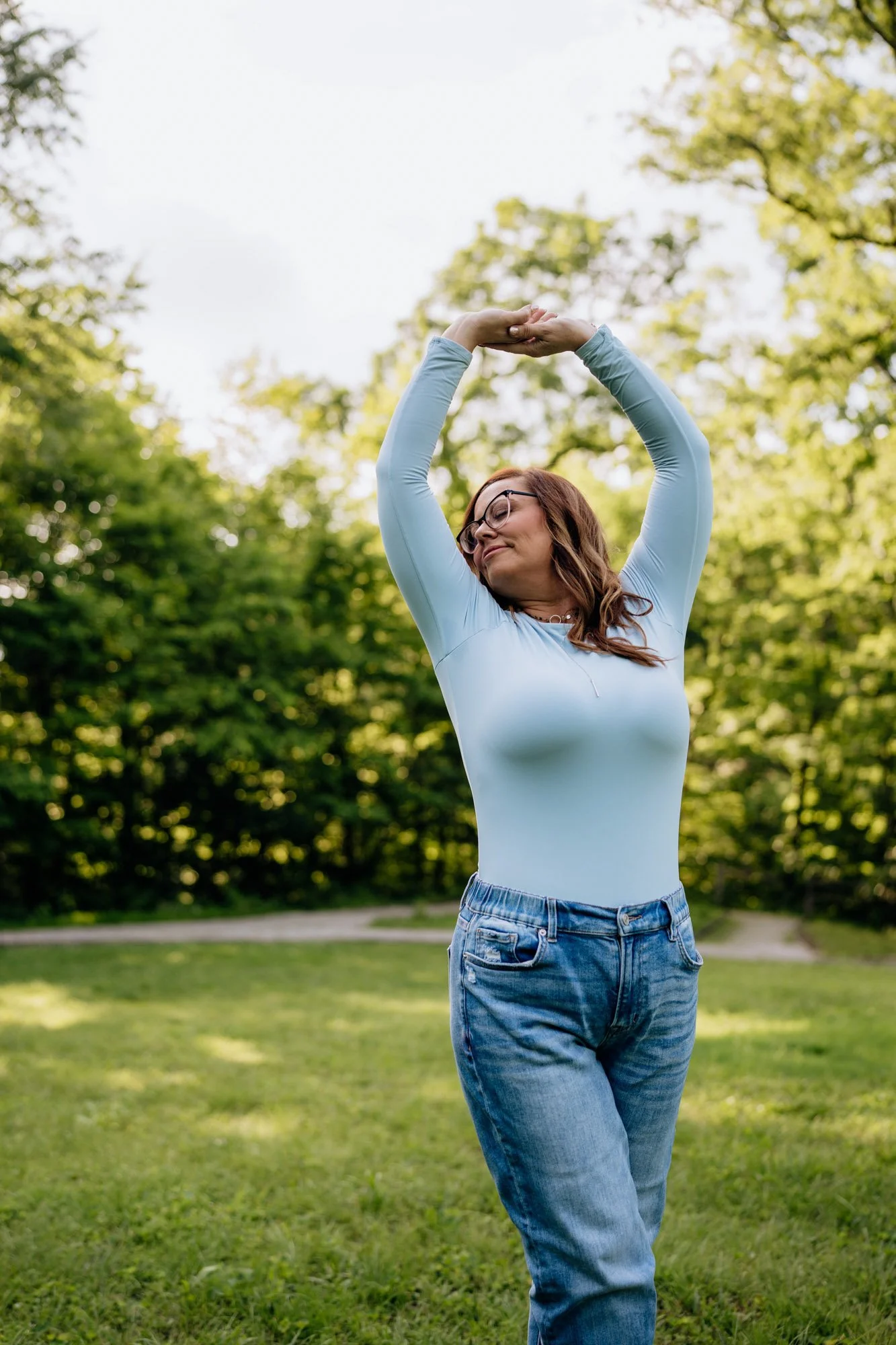 A woman in a light blue long sleeve shirt and jeans doing a stretch outdoors in a park with green trees.