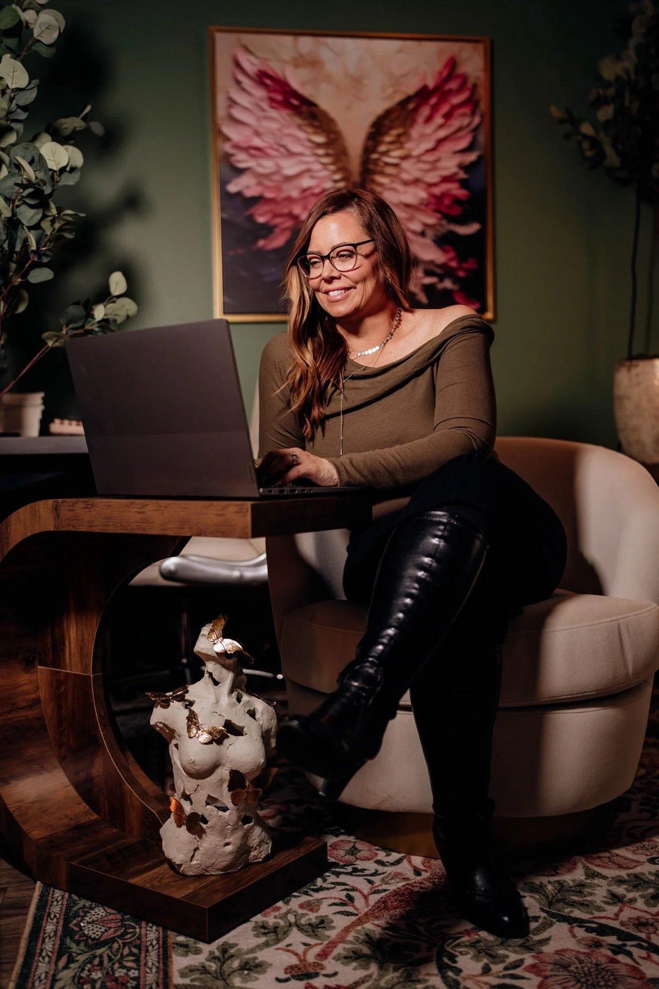 A woman with long brown hair and glasses sitting on a beige armchair, smiling at her laptop in a room with green walls. Behind her is a framed picture of angel wings and large green plants. She is wearing an off-shoulder brown top and black leather pants, with a decorative ceramic angel sculpture on a wooden side table in front of her and a patterned rug on the floor.