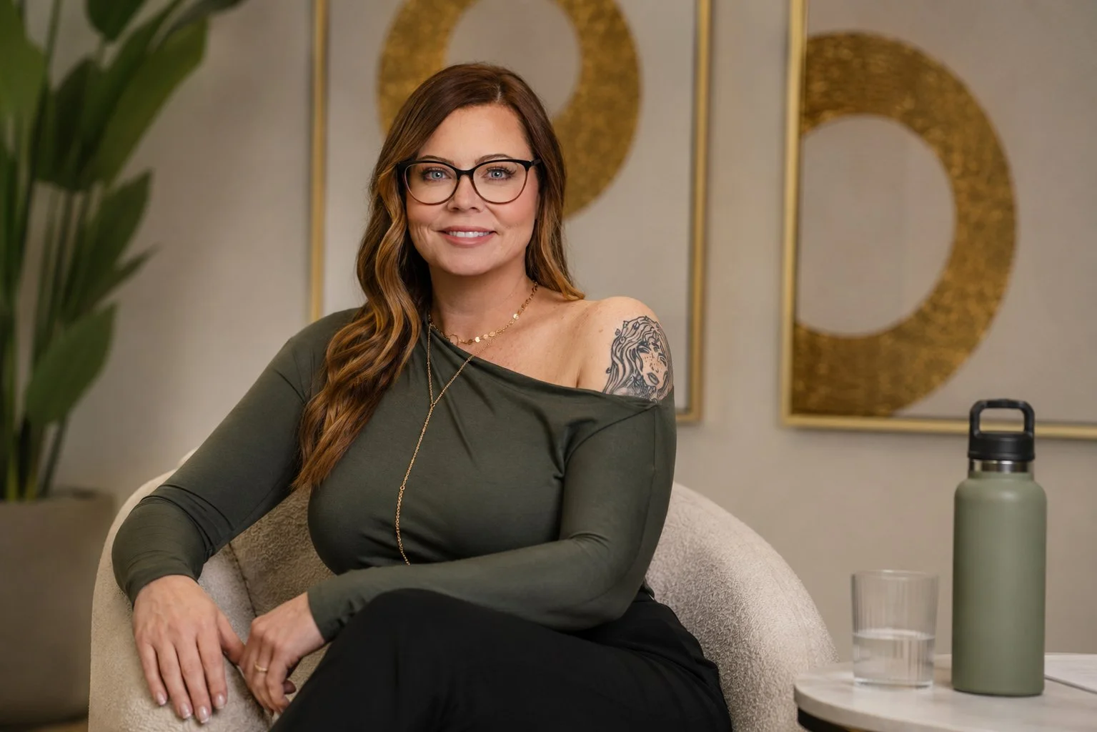 A woman with glasses and long wavy brown hair sits on a beige armchair, smiling at the camera. She wears an olive green off-the-shoulder top, revealing a tattoo on her right shoulder, and layered gold necklaces. In the background, there are framed gold and white art pieces, a leafy plant on the left, and a small table on the right with a thermos and a glass of water.