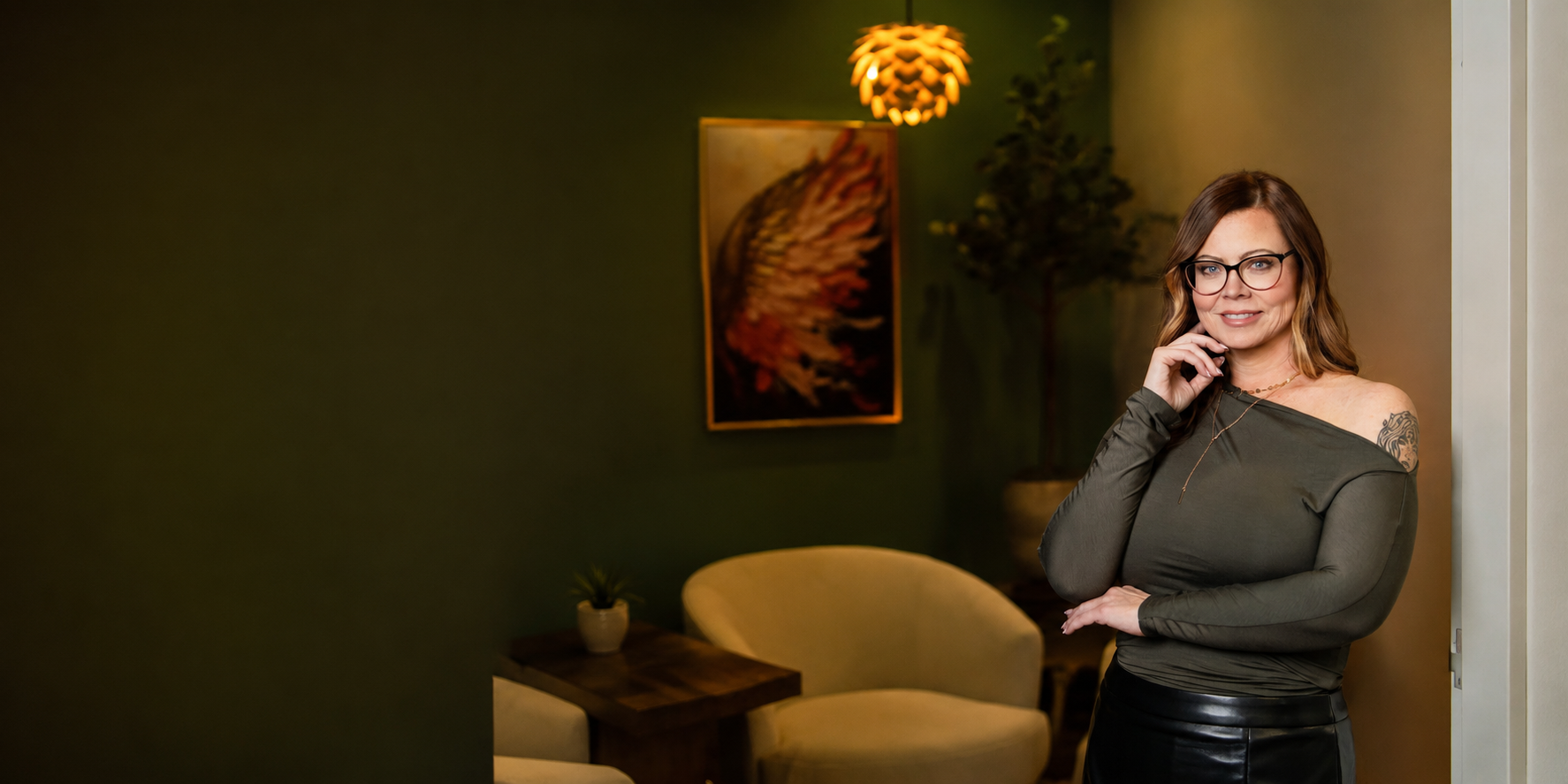 A woman with glasses and wavy hair standing in a room with green walls, artwork, and furniture, looking at the camera with a smile.
