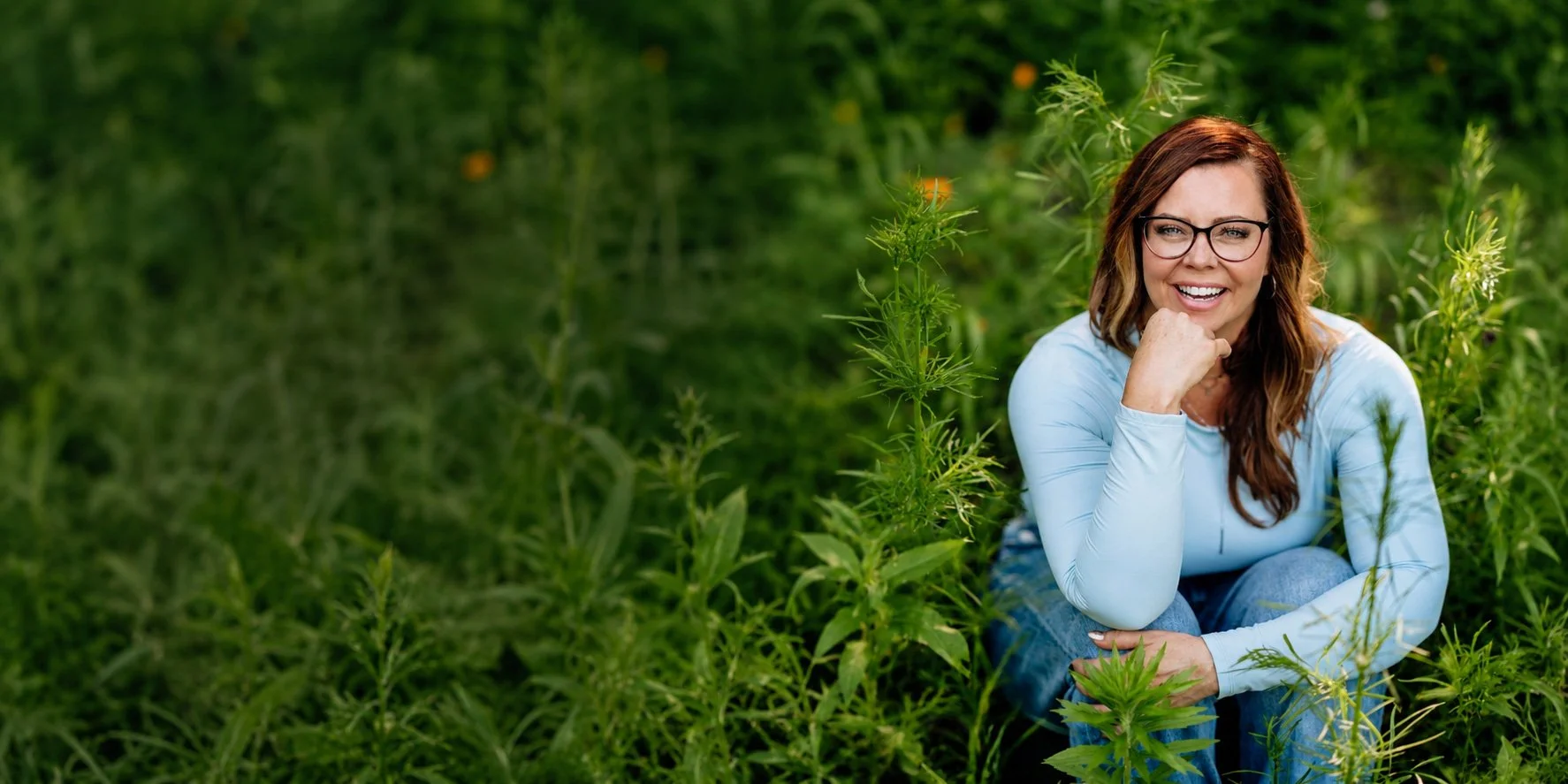 A woman with red hair and glasses sitting in a green garden, smiling, wearing a light blue long-sleeve shirt and jeans.