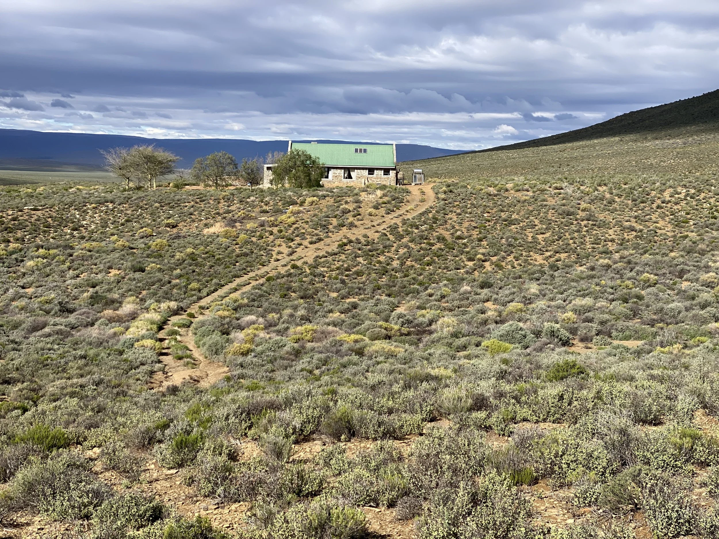 A small stone building with a green metal roof situated in a desert landscape with sparse vegetation, trees, and distant mountains under a cloudy sky.