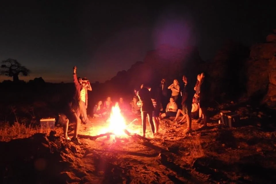 Group of people gathered around a campfire outdoors at night, some sitting and some standing.