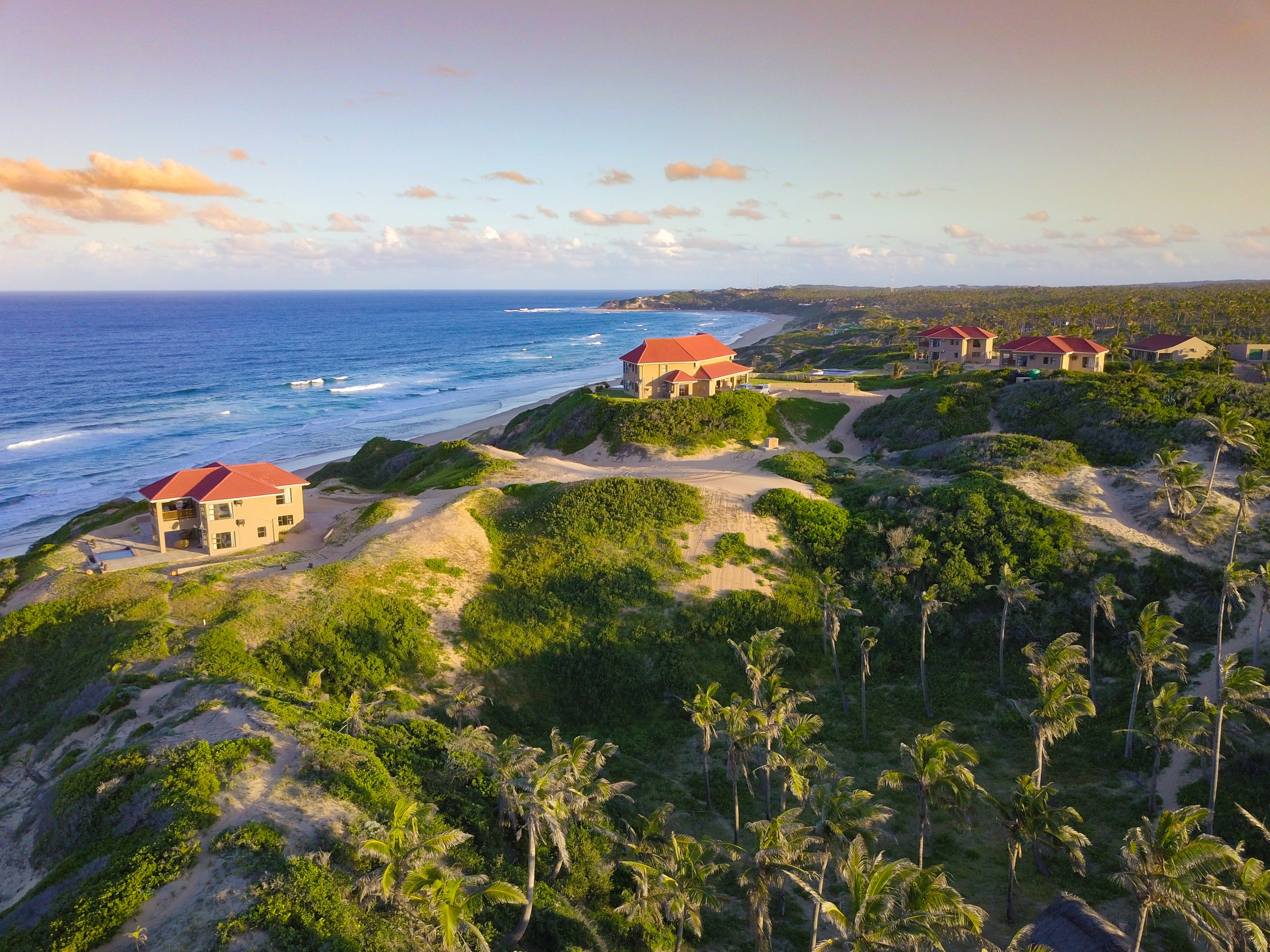 Aerial view of a coastal area with sandy beaches, ocean waves, and several houses with red roofs among lush green vegetation.