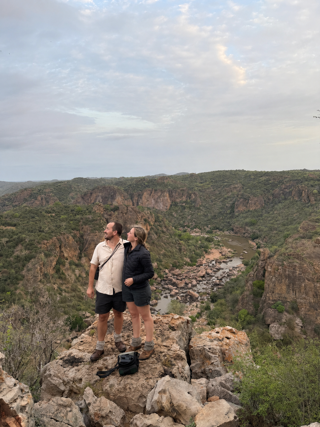 A couple standing on a large rock overlooking a canyon with a river, surrounded by green hills and a cloudy sky.