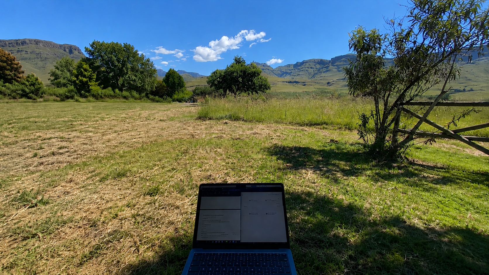 A laptop on grass in a field with trees, mountains, and a blue sky with clouds in the background.