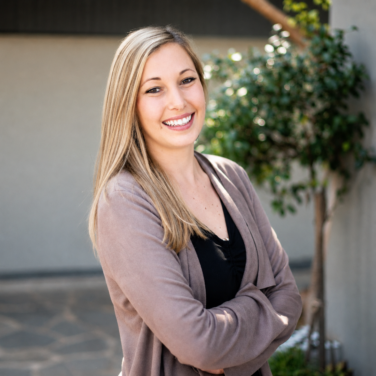 A young woman with long blonde hair smiling and standing outdoors with crossed arms, wearing a light brown blazer over a black top, and a background of a gray wall, tree, and greenery.