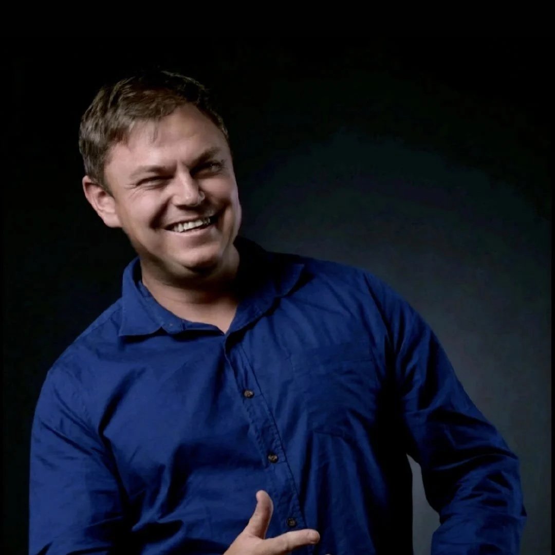 A man with short brown hair wearing a blue button-up shirt, smiling and giving a thumbs-up gesture against a dark background.