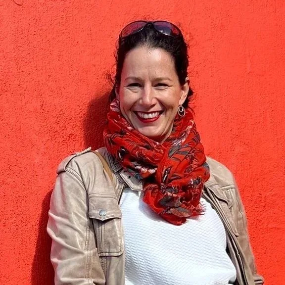 A woman smiling in front of a bright orange wall, wearing sunglasses on her head, a red patterned scarf, a beige jacket, and a white shirt.