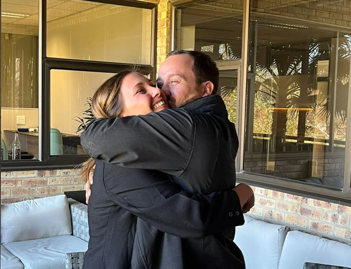 A man and woman hugging indoors, with large windows showing plants outside.