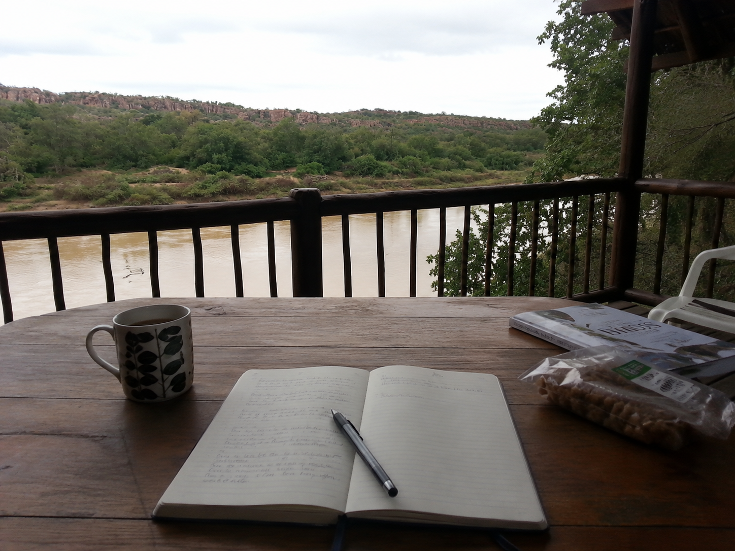 A wooden table on a balcony overlooking a river, with a notebook, pen, mug, reader, and snack on the table. Green trees and hills are in the background, under a cloudy sky.