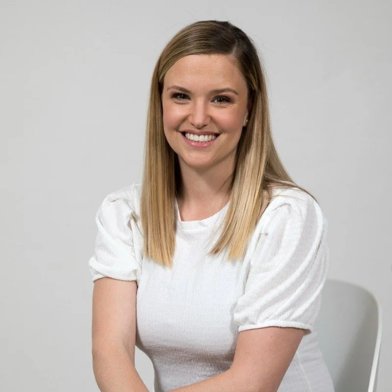 A young woman with straight blonde hair, smiling, wearing a white short-sleeved top, sitting against a plain white background.