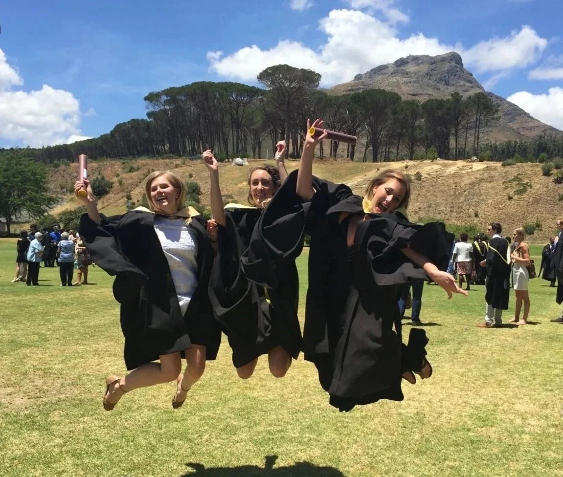 Three women in graduation gowns jumping in the air on a grassy field during a graduation ceremony, with a mountain and trees in the background.
