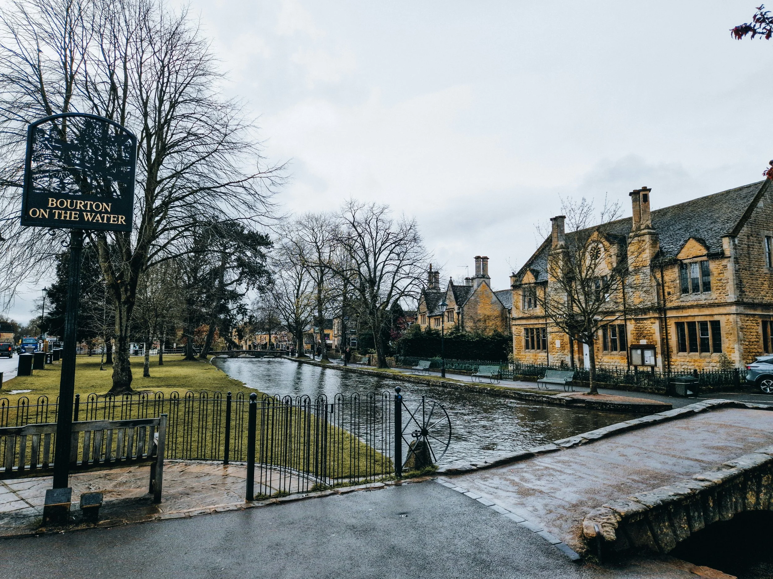 A park in Bourton-on-the-Water with a small stream, benches, trees, and historic stone buildings with chimneys on a cloudy day.